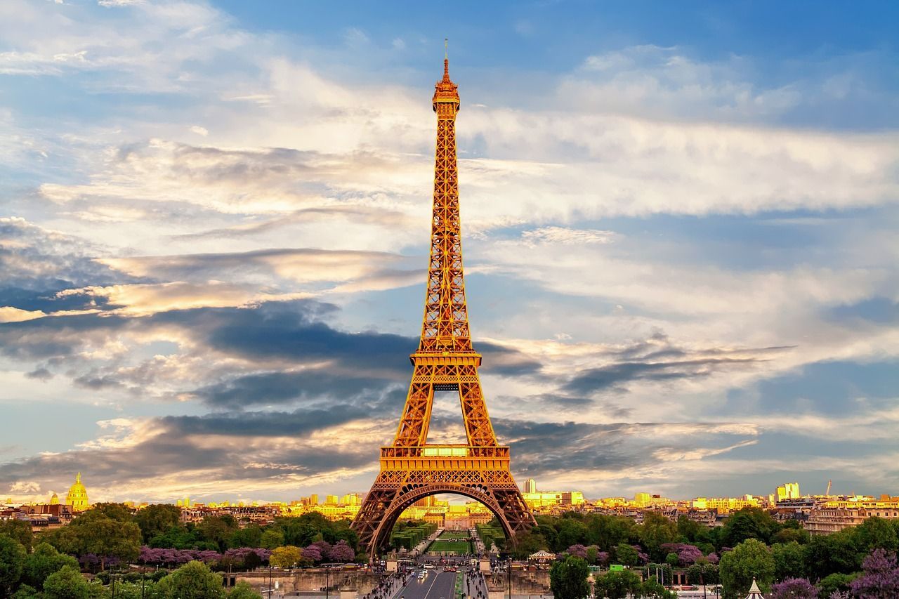 La Torre Eiffel si erge imponente contro un cielo parzialmente nuvoloso al tramonto, con lo skyline di Parigi e alberi verdi sottostanti.