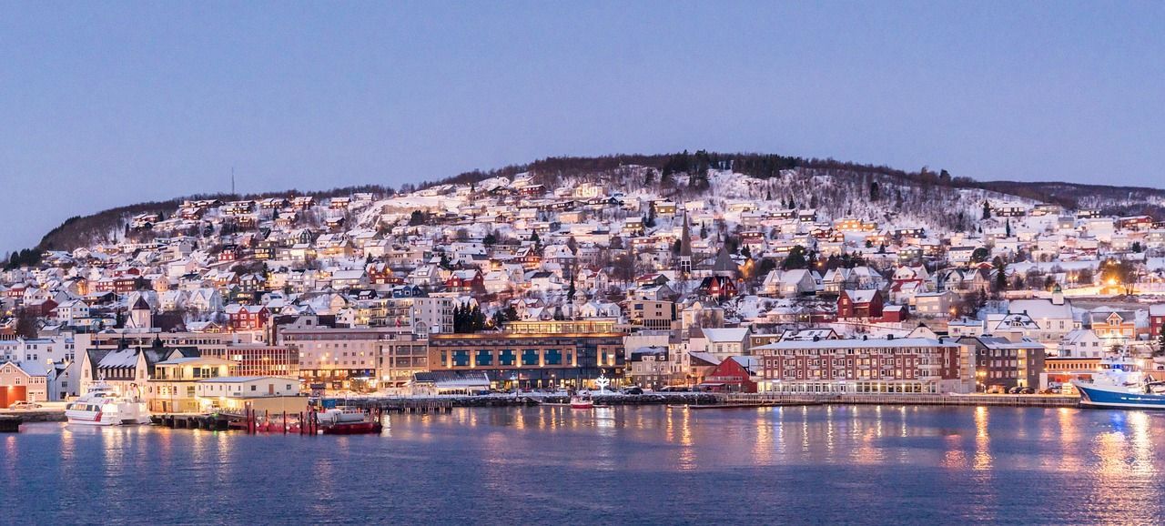 Una città costiera con edifici illuminati su un pendio innevato, vista dall'acqua al tramonto con le luci che si riflettono sulla superficie.