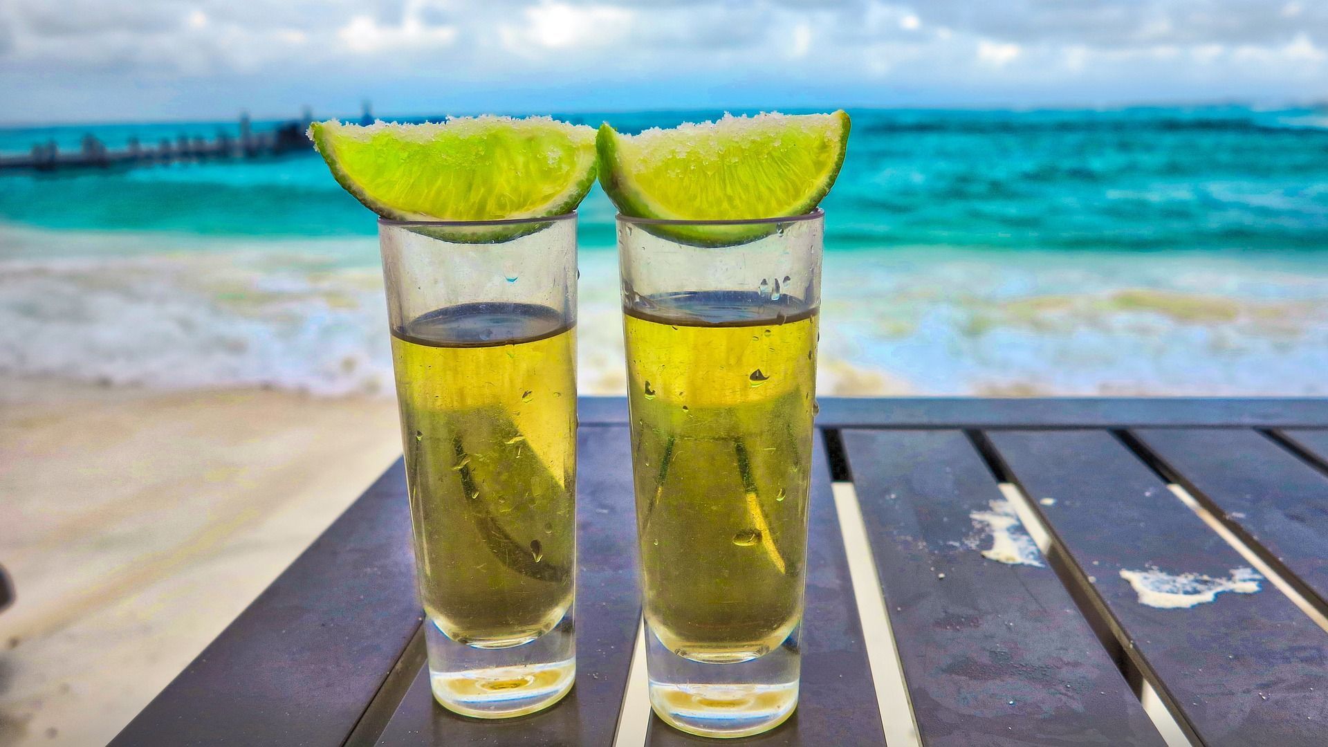 Due shot di tequila con spicchi di lime salati poggiano su un tavolo con vista su un oceano turchese e una spiaggia sabbiosa.