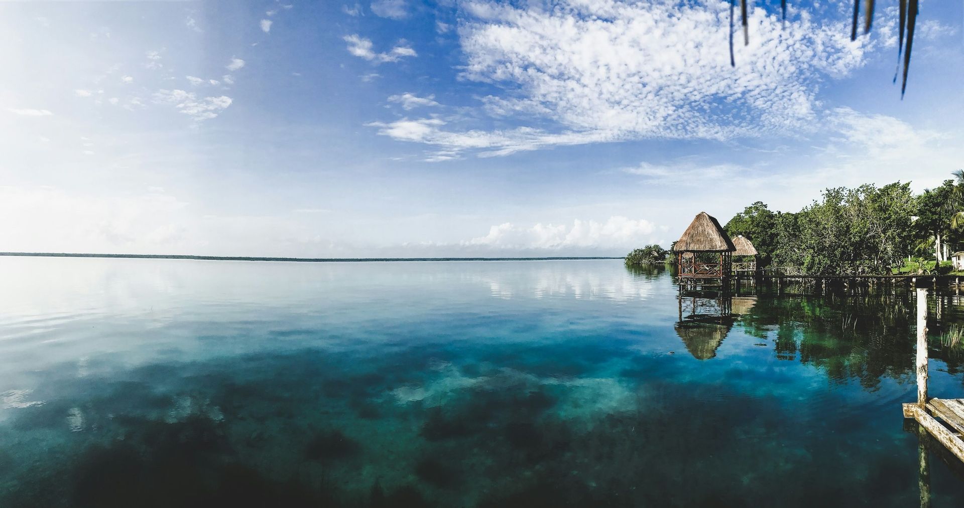 Bungalow dal tetto di paglia sulla riva di una laguna tranquilla si riflettono nell'acqua calma e turchese sotto un cielo blu.