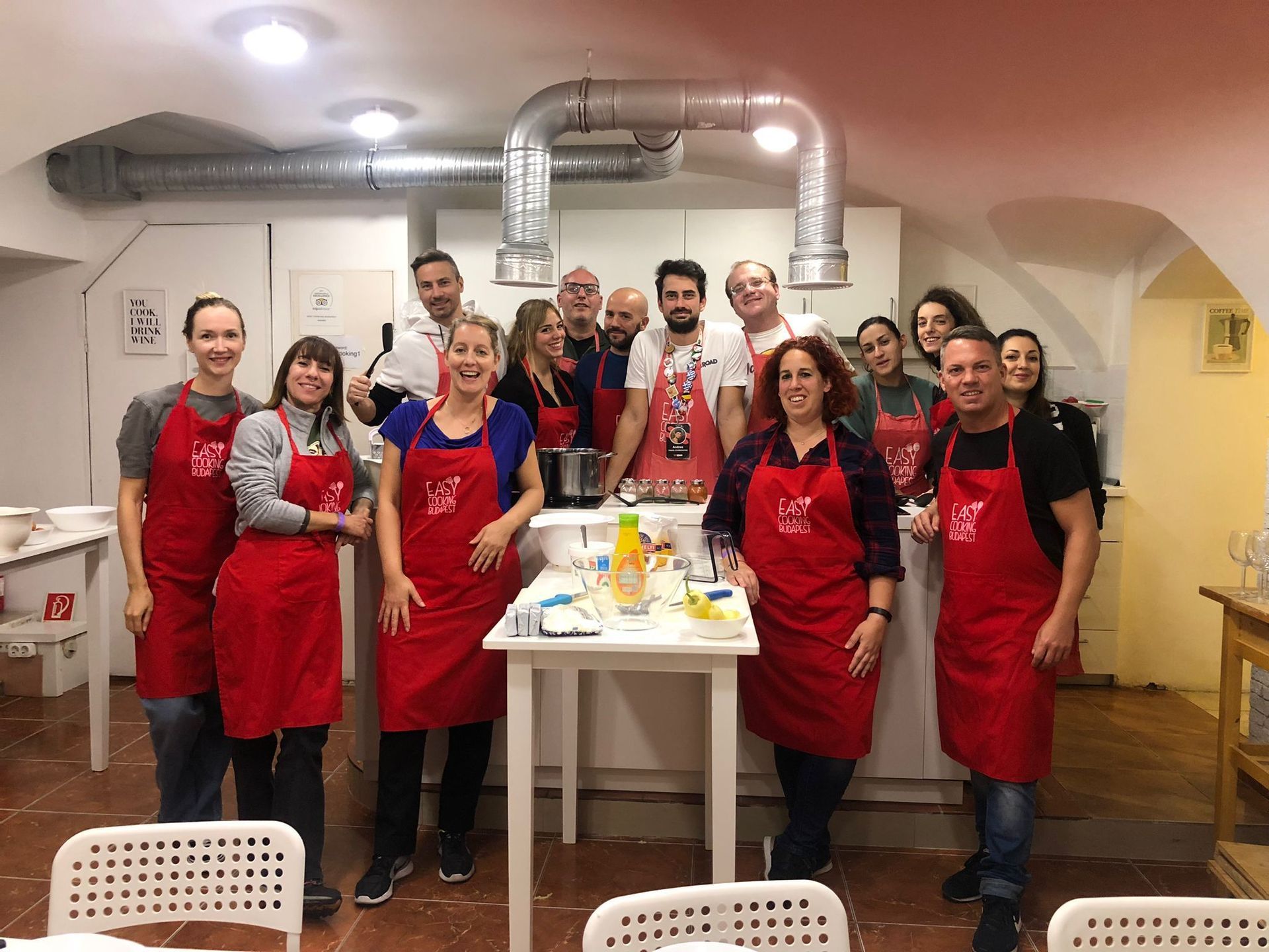 Un gruppo WeRoad sorride per una foto durante una lezione di cucina, tutti indossano grembiuli rossi coordinati in una cucina.