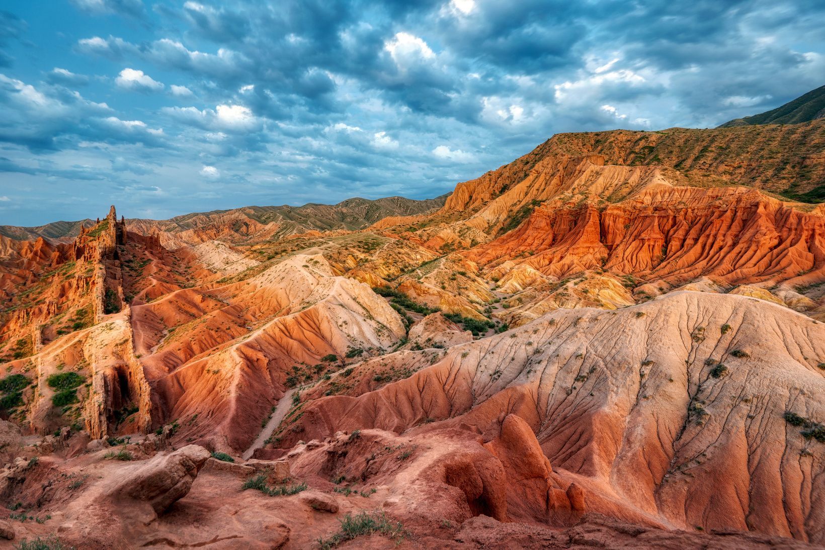 Eine weite Ansicht einer trockenen Landschaft mit roten und orangefarbenen Canyon-Formationen unter einem bewölkten blauen Himmel.