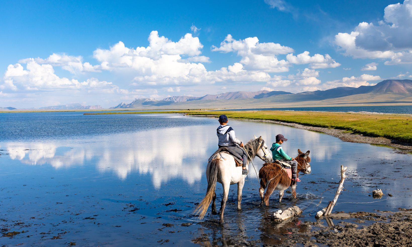 Deux personnes à cheval et à dos d'âne traversent un lac paisible reflétant les nuages et les montagnes lointaines.