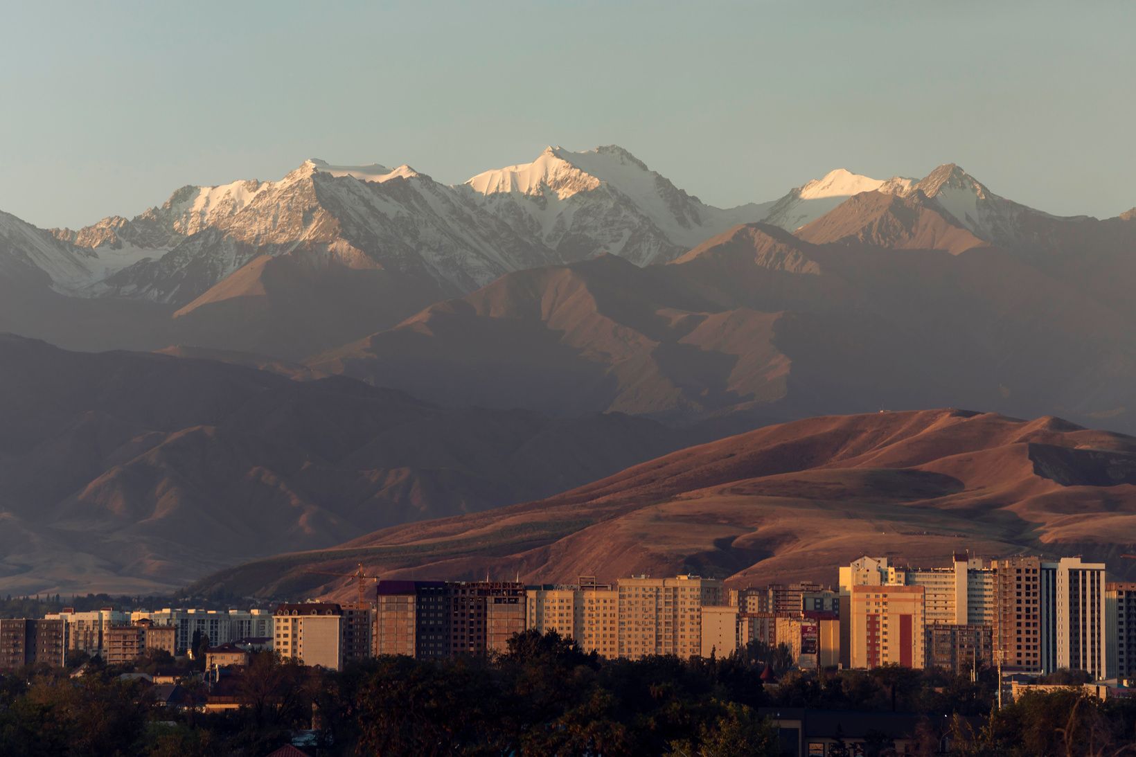 Eine Stadt-Skyline mit Gebäuden liegt am Fuße sanfter Hügel, im Hintergrund eine große, schneebedeckte Bergkette.