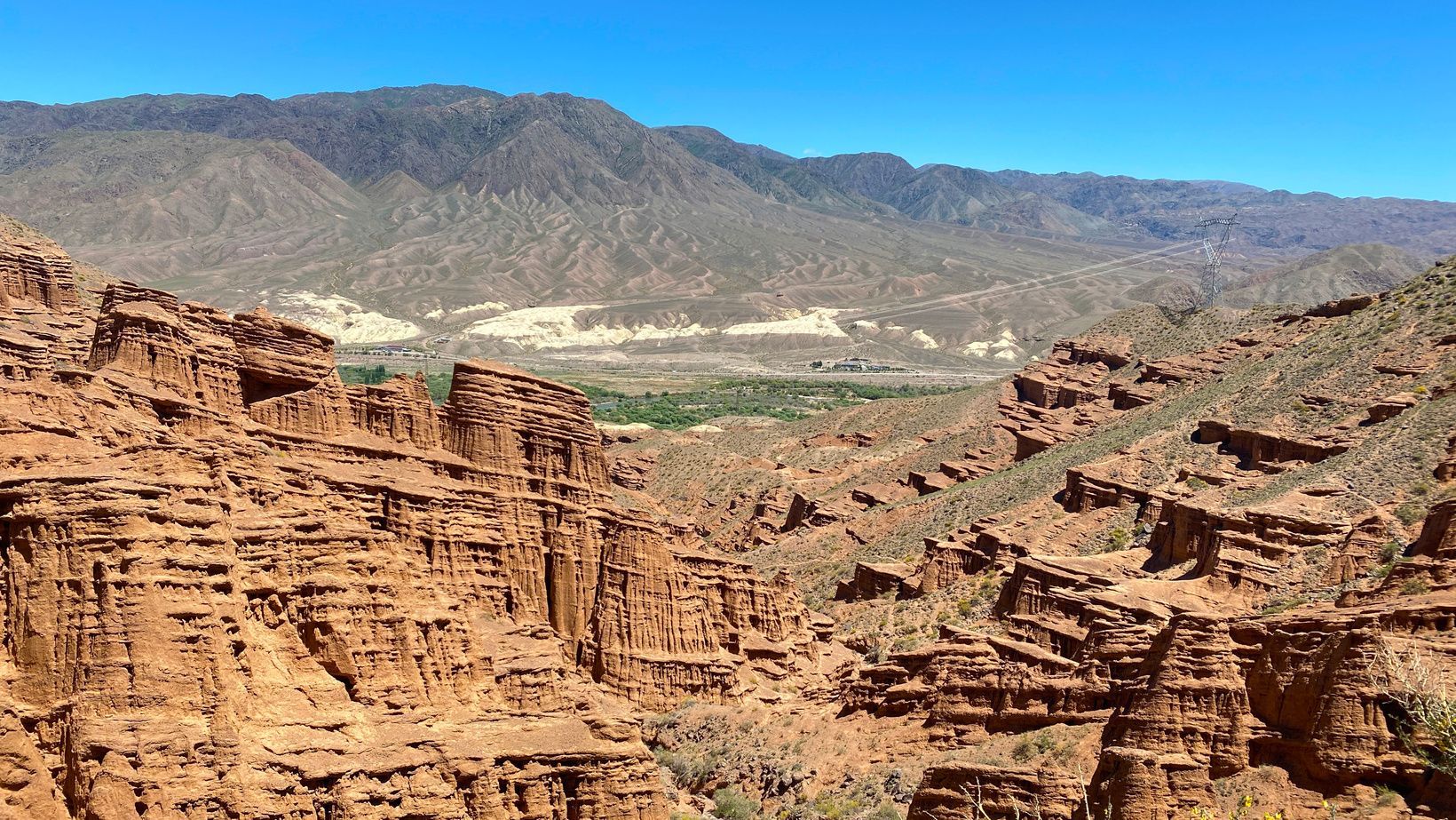Una vista su un canyon con formazioni rocciose rosse stratificate, con montagne aride in lontananza sotto un cielo azzurro brillante.
