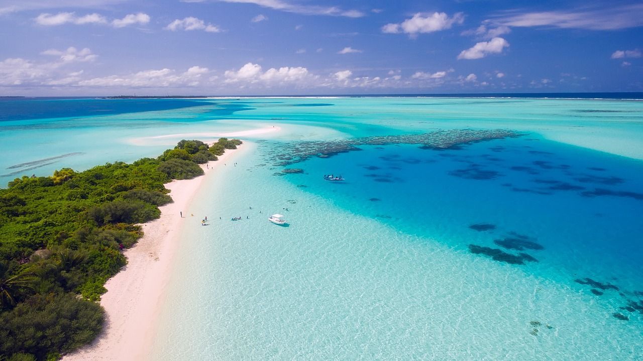 Una vista aerea di una lingua di sabbia tropicale con una spiaggia bianca, barriera corallina e barche in acqua cristallina turchese.