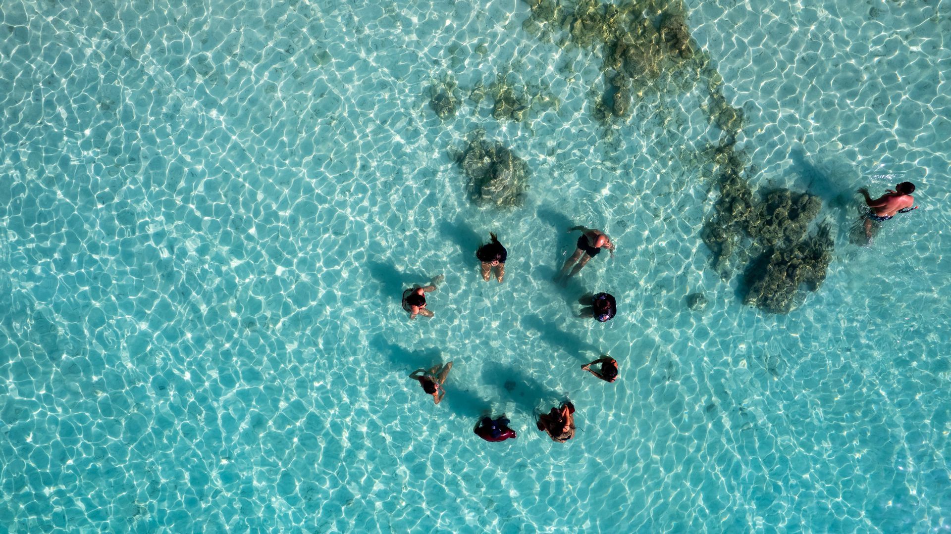 Vista aerea di un gruppo WeRoad in cerchio in acqua bassa, limpida e turchese.