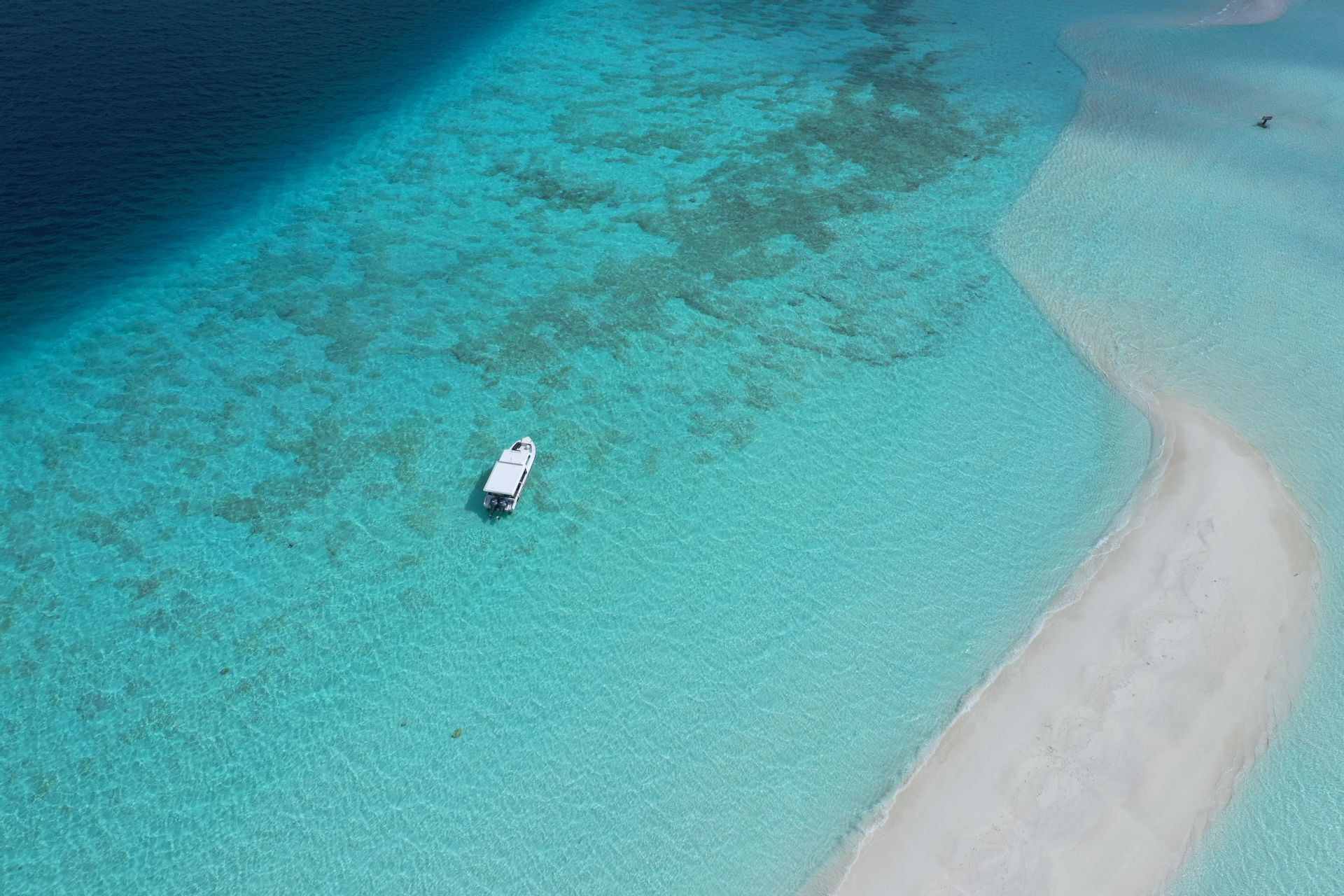 Una vista aerea di una barca bianca in acqua turchese accanto a una lingua di sabbia bianca, in contrasto con il mare blu profondo.