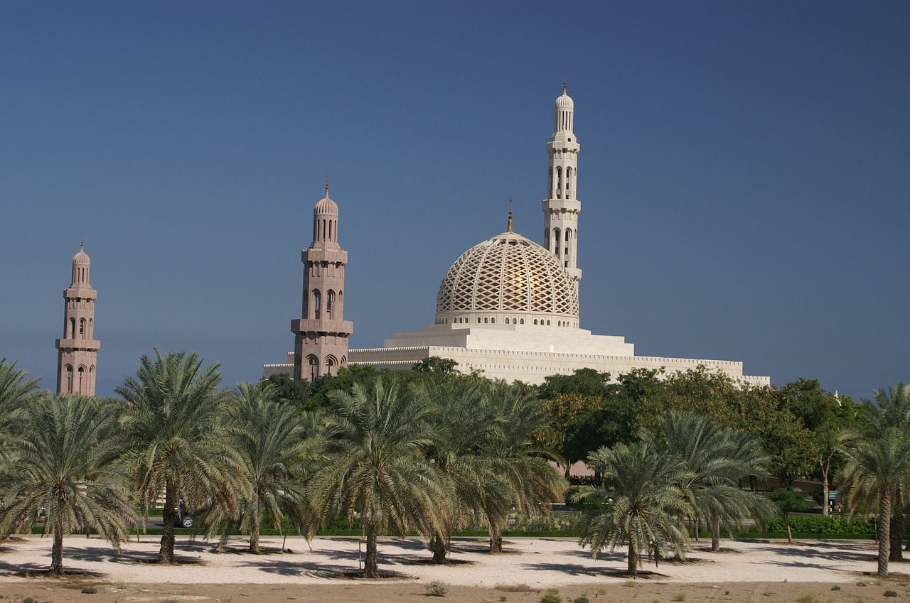 Una grande moschea con una cupola dorata a griglia e alti minareti si erge dietro un palmeto sotto un cielo azzurro limpido.