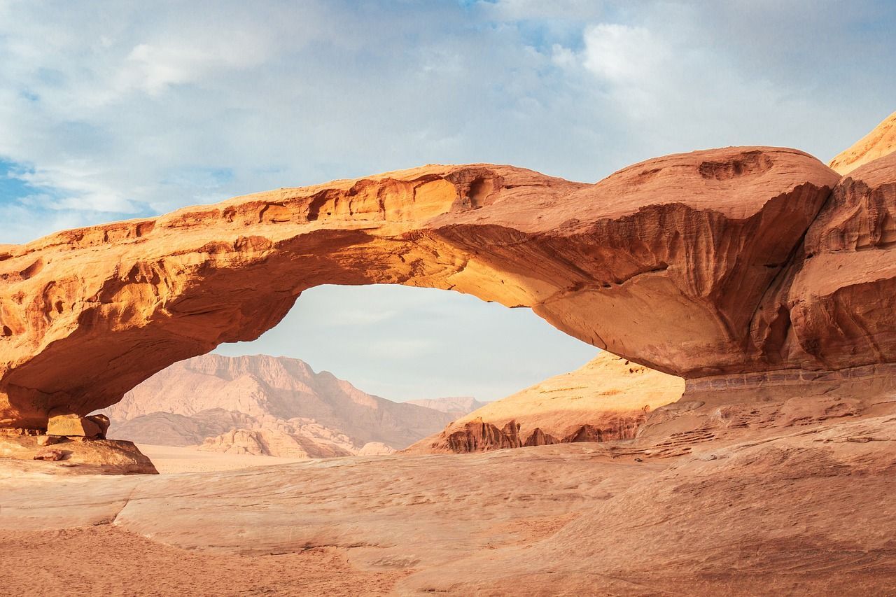 Un arco naturale di arenaria incornicia montagne lontane in un paesaggio desertico sotto un cielo parzialmente nuvoloso.