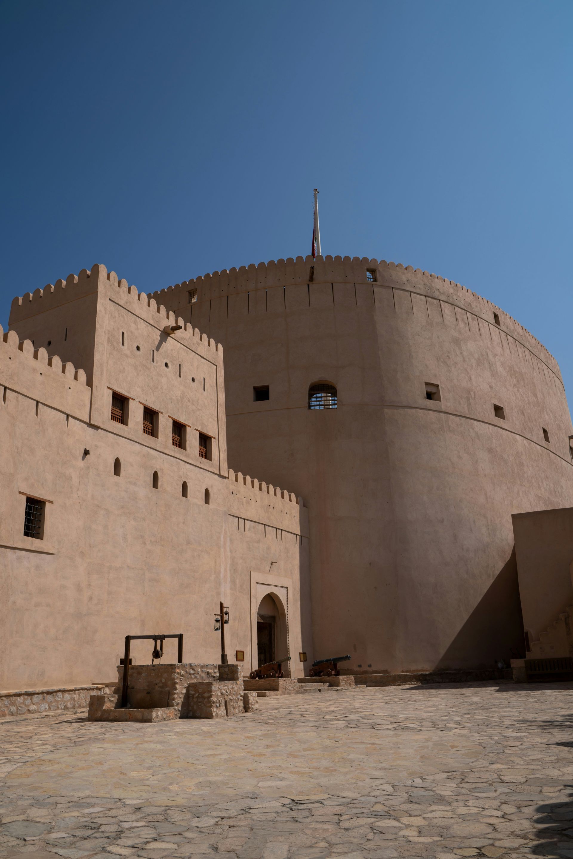 Un imponente forte color sabbia con una torre rotonda domina un cortile in pietra contenente un pozzo e cannoni, sotto un cielo blu limpido.