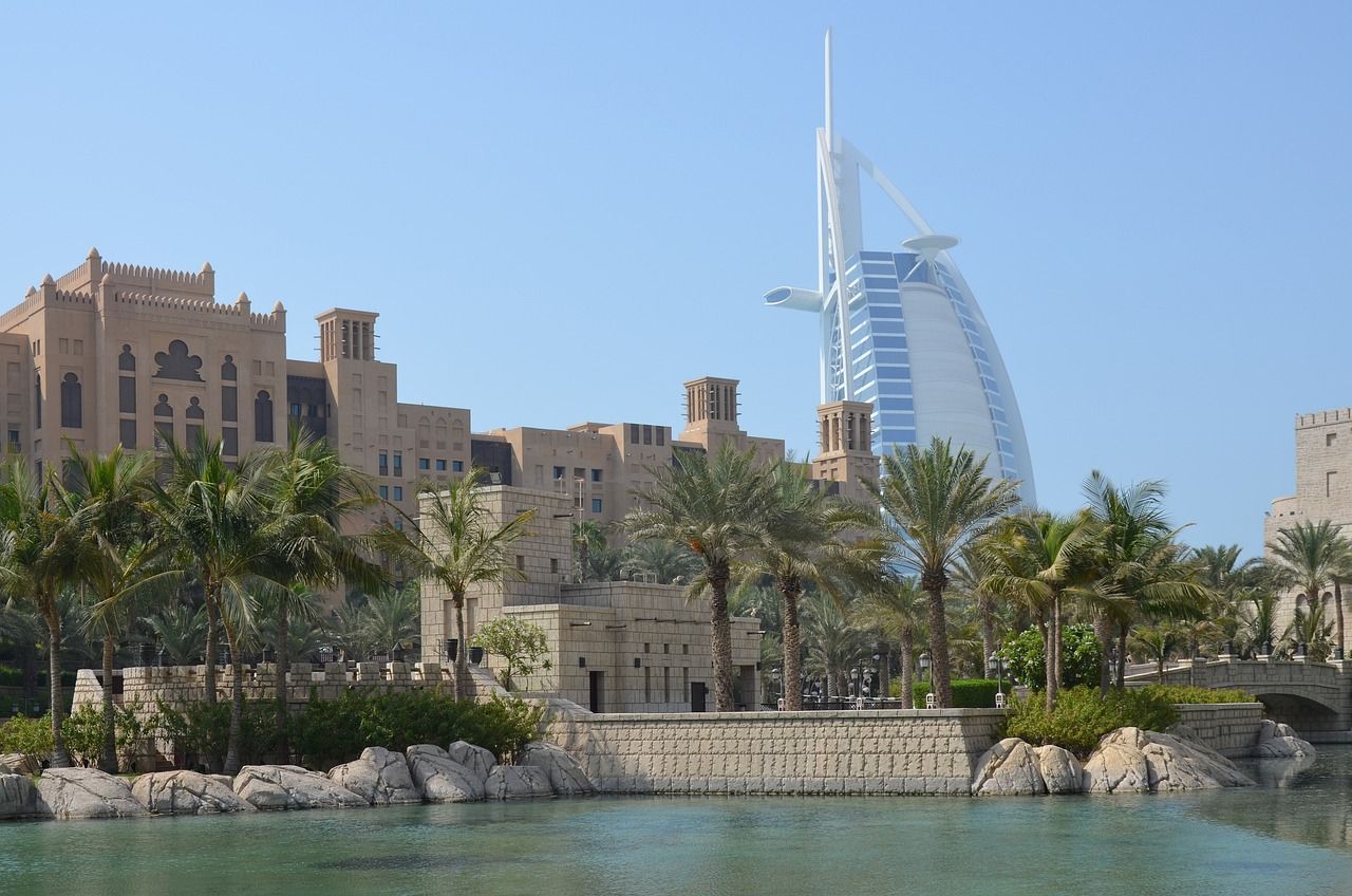 Un grattacielo a forma di vela si erge dietro edifici tradizionali color sabbia e palme, con uno specchio d'acqua calmo in primo piano.