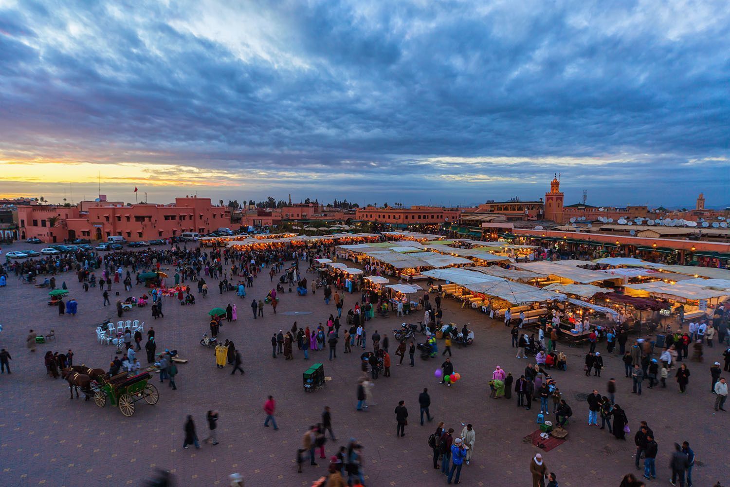 Una vista elevada de una bulliciosa plaza de ciudad abarrotada de gente y puestos de mercado al atardecer, con edificios de terracota al fondo.