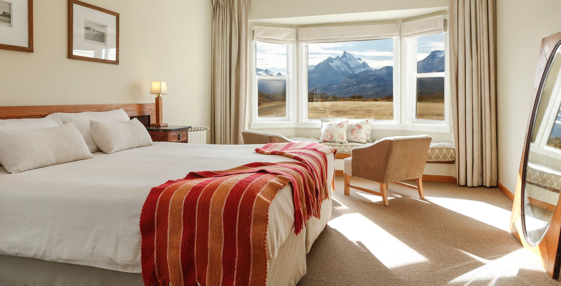 Una camera da letto illuminata dal sole con un letto fatto e un plaid a righe, dotata di una finestra a bovindo con vista su montagne innevate.