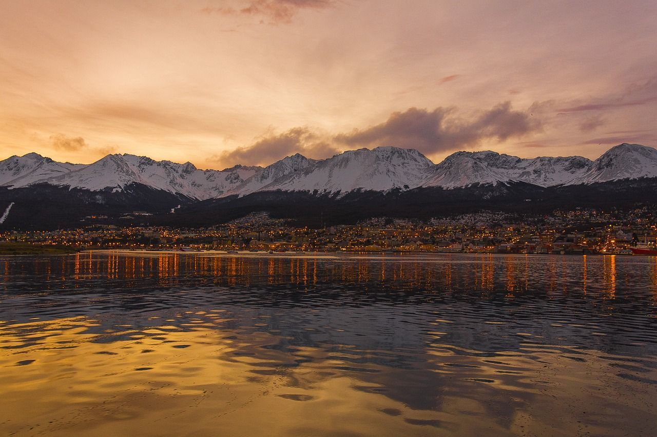 Le luci di una città costiera si riflettono sull'acqua al tramonto, con una catena di montagne innevate sullo sfondo.