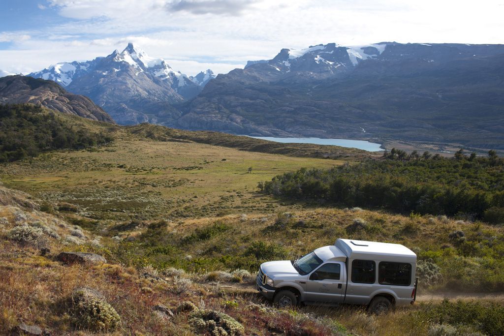 Un furgone argentato percorre una strada sterrata attraverso un paesaggio collinare con un lago e montagne innevate in lontananza.