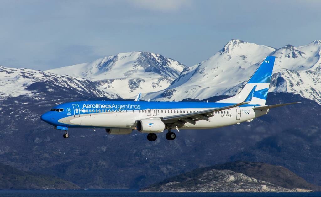 Un aereo di Aerolíneas Argentinas vola con il carrello abbassato davanti a una catena di montagne innevate.