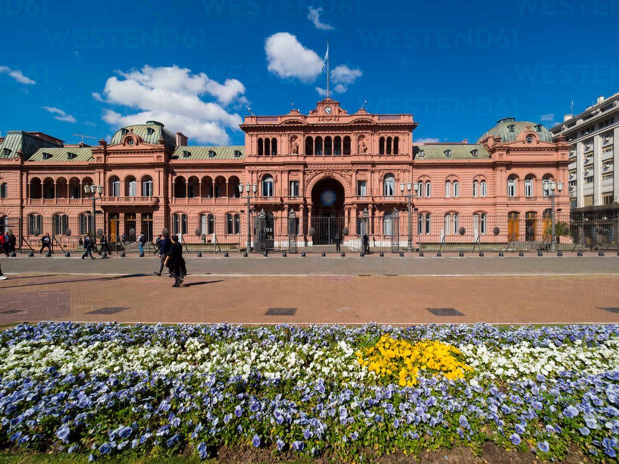 Un grande edificio governativo rosa con una bandiera, visto da una piazza con un'aiuola colorata in primo piano in una giornata di sole.