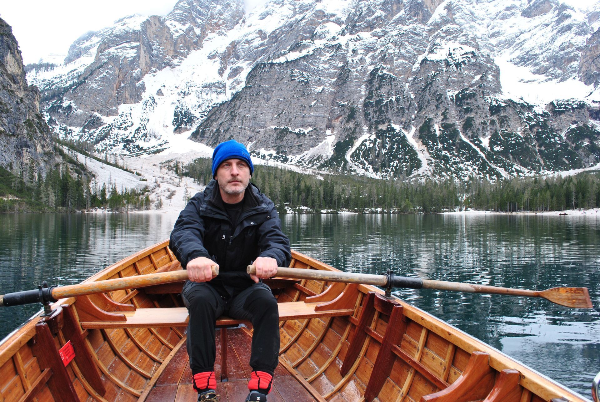 Un uomo con un berretto blu rema una barca di legno su un lago con montagne innevate sullo sfondo.