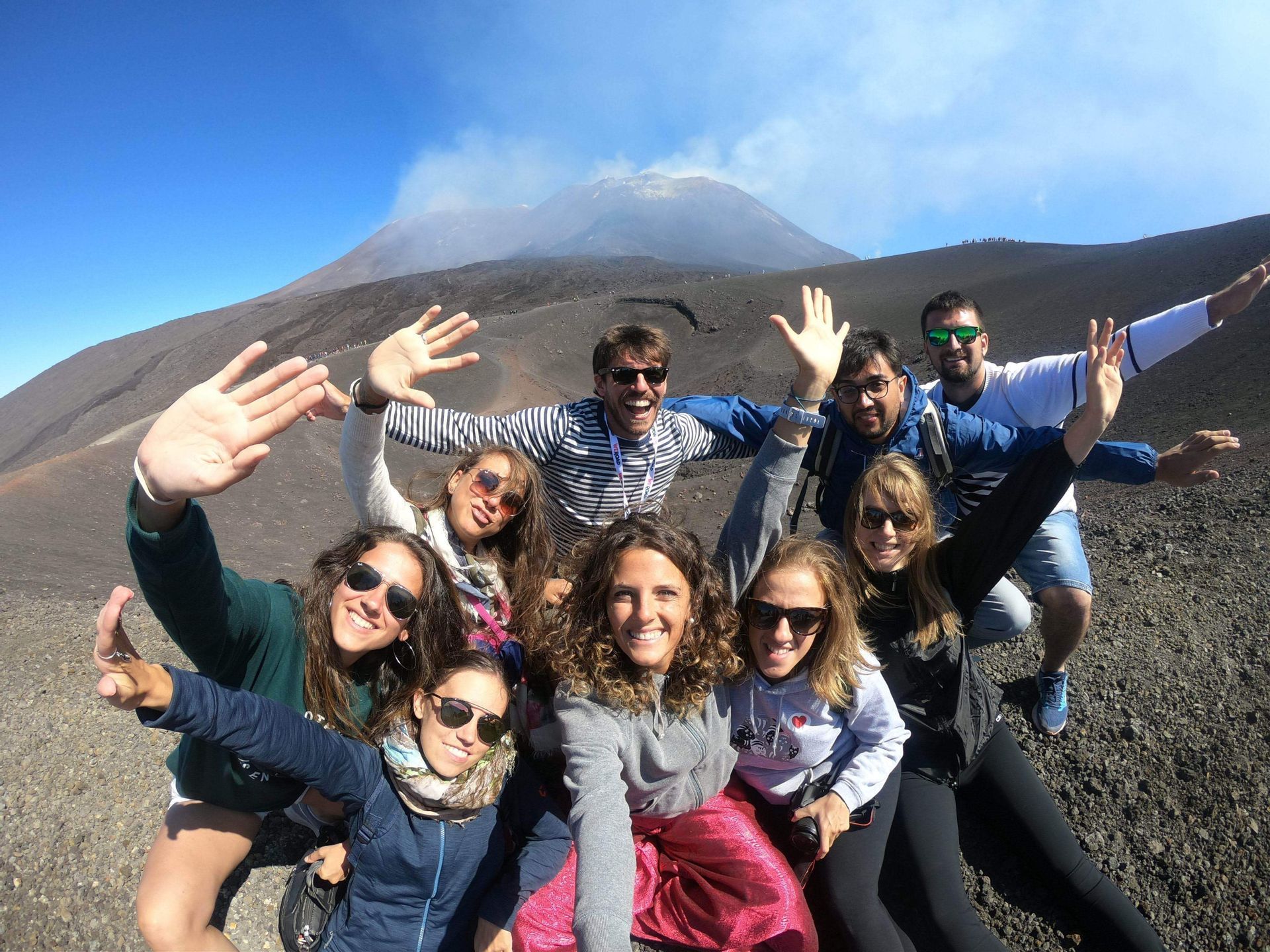 Un grupo de WeRoad se toma un selfi en una ladera rocosa, con un volcán humeante al fondo bajo un cielo azul claro.