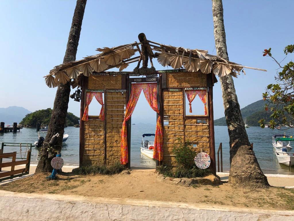 A rustic mud-brick gateway with a thatched roof stands between two palm trees on a beach, framing a view of the sea with a boat.