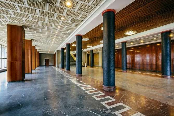 Una lunga sala con ampie colonne nere, un pavimento in marmo lucido con motivo geometrico e pareti rivestite in legno.