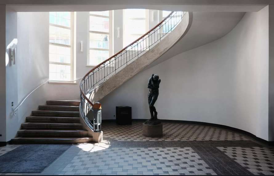 Una sala illuminata dal sole con una scala curva in cemento, una scultura scura su un piedistallo e un pavimento a scacchi.