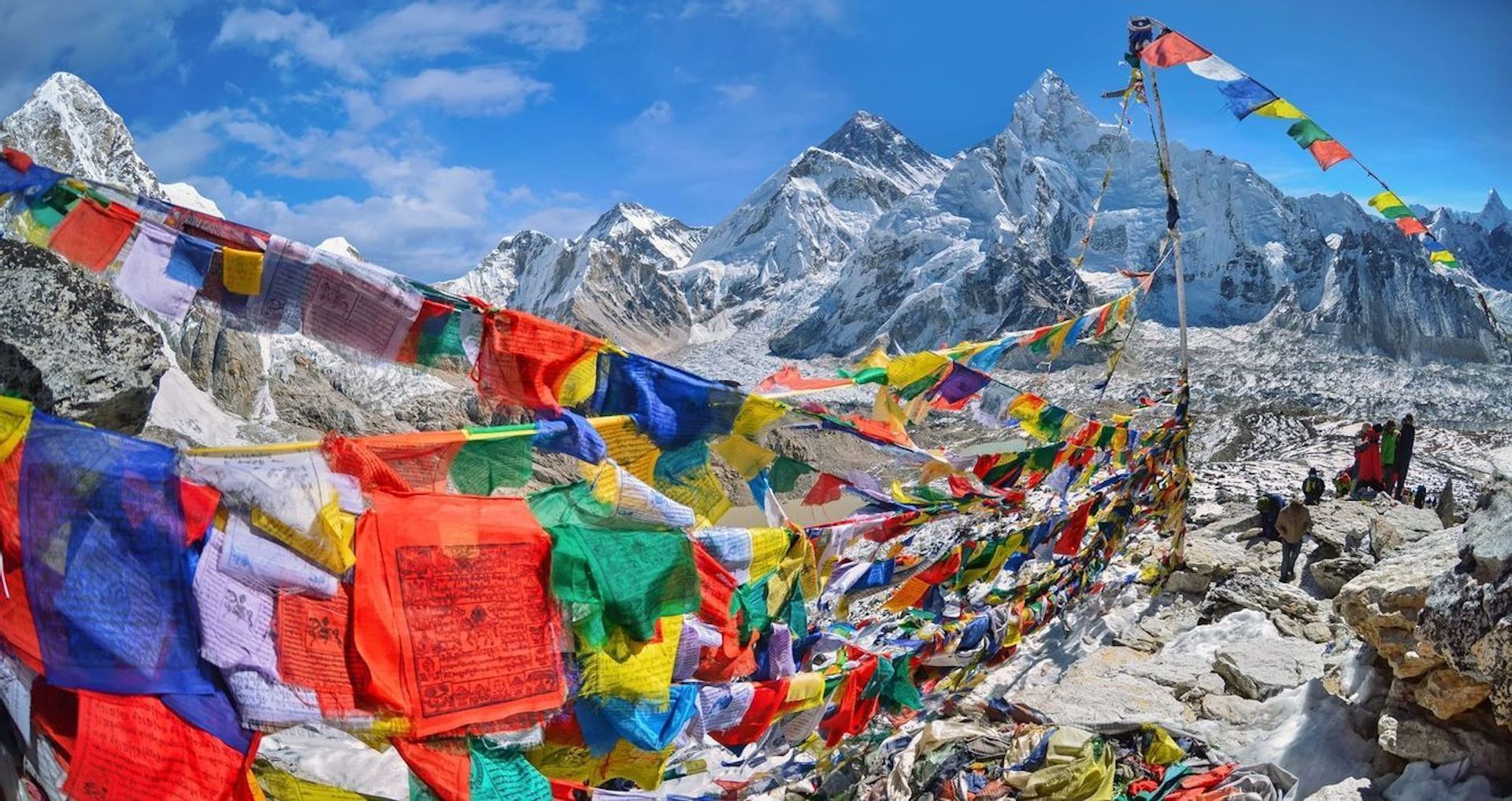 Un viaje en grupo de WeRoad contempla montañas nevadas desde un mirador rocoso adornado con coloridas banderas de oración bajo un cielo azul.