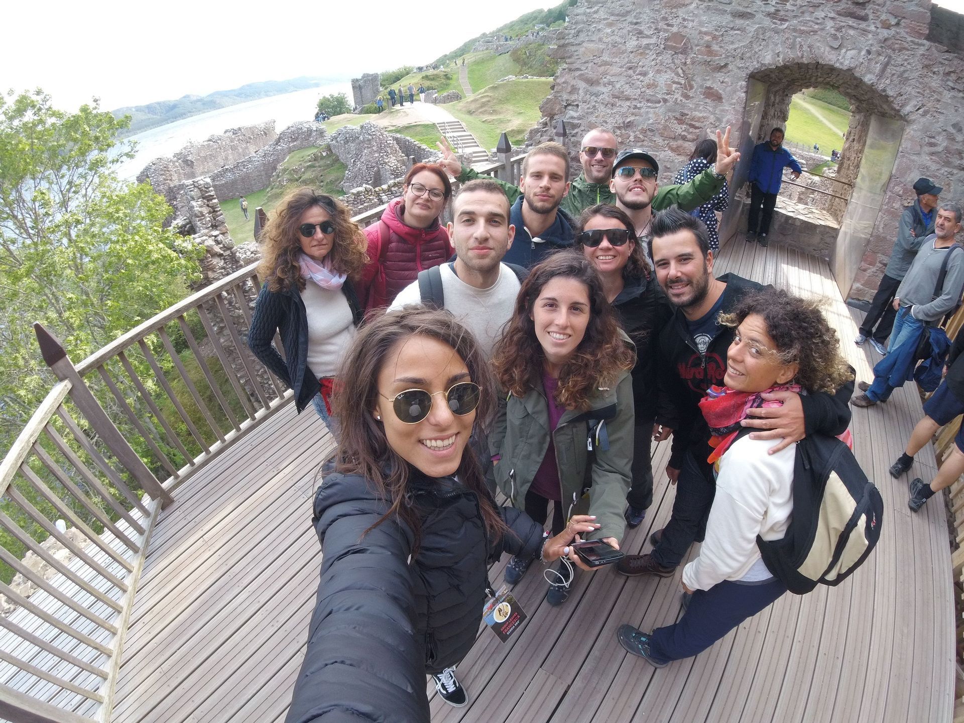 A WeRoad group trip takes a smiling selfie on a wooden walkway with castle ruins and a body of water in the background.