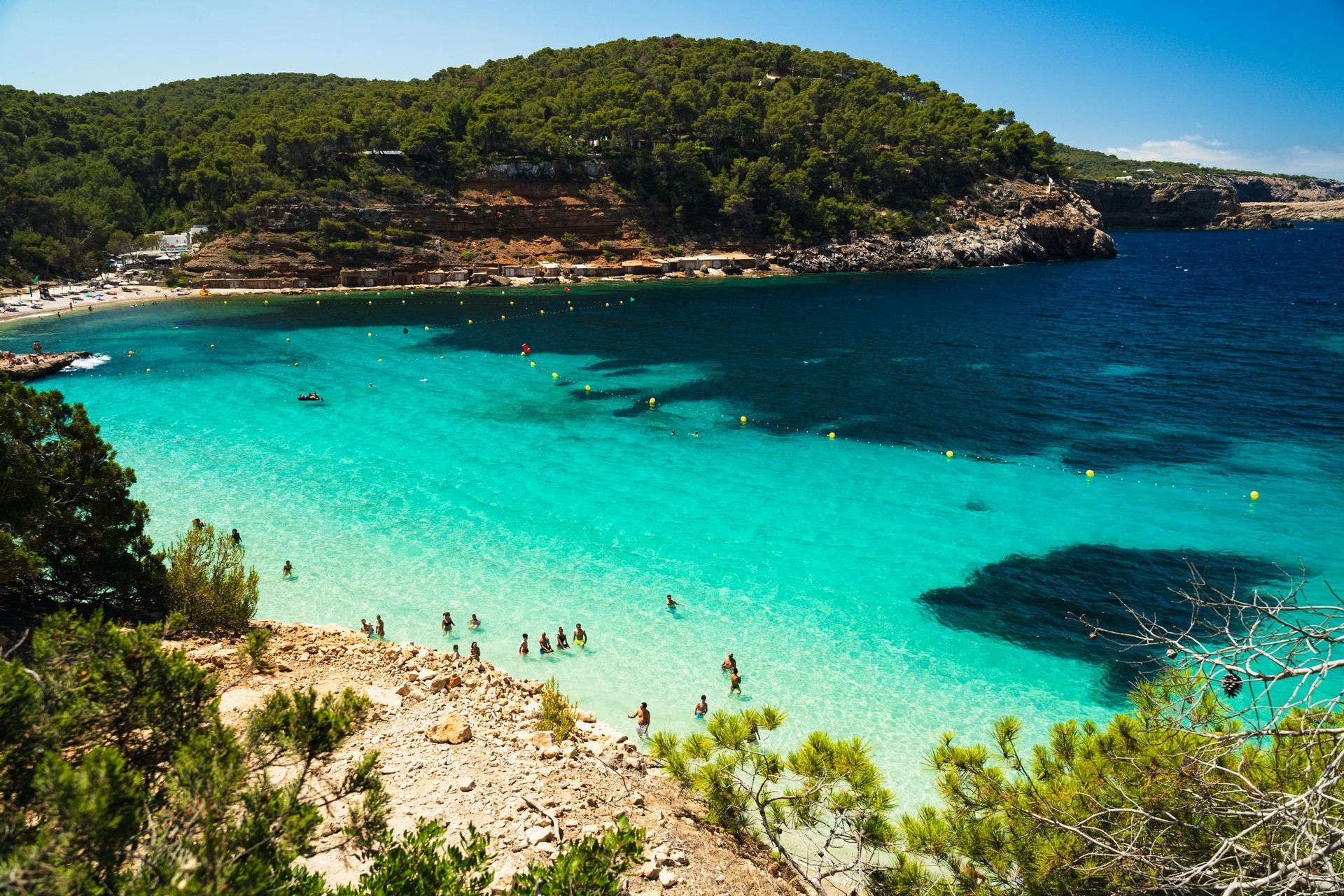 Una vista dall'alto di una cala dove le persone nuotano in acque turchesi poco profonde, con una spiaggia e una scogliera boscosa sullo sfondo.