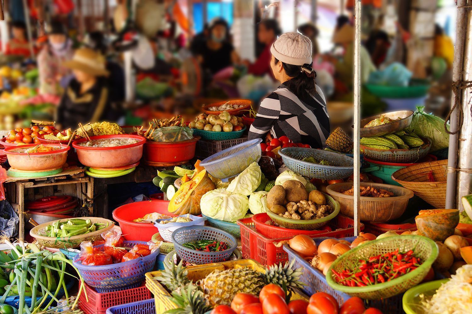 Eine Frau an einem belebten Freiluftmarktstand, prall gefüllt mit frischem Gemüse, Obst und weiteren Produkten in farbenfrohen Körben.