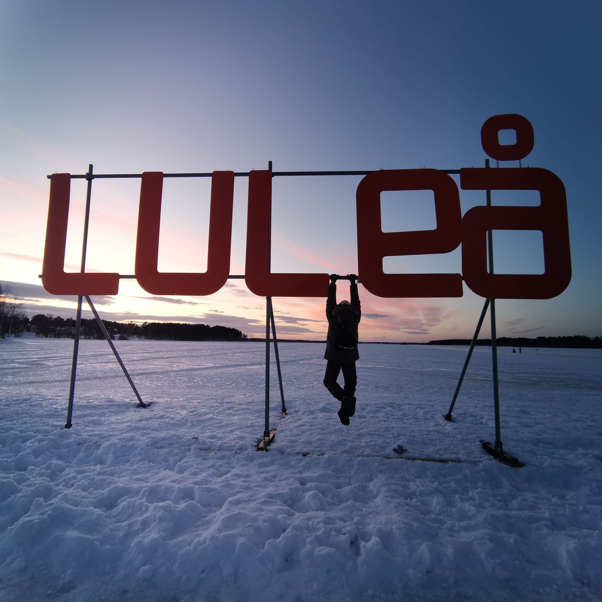 Una persona in controluce appesa alla grande insegna rossa 'LULEÅ' in un paesaggio innevato al tramonto.