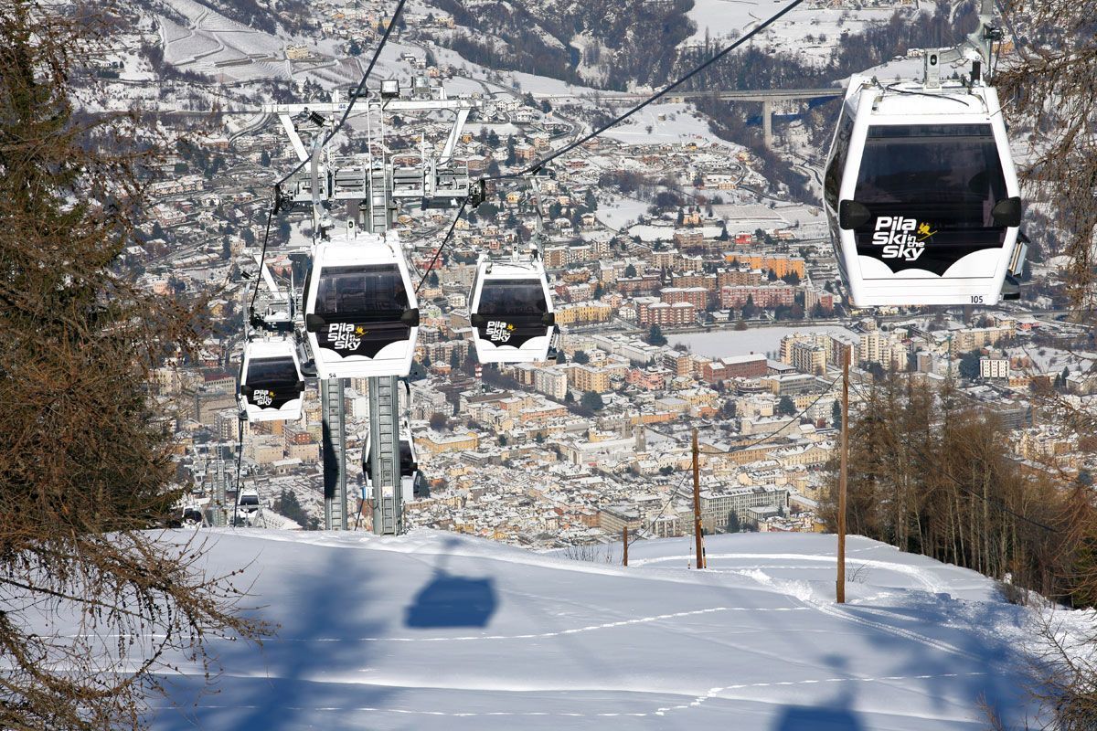 Una fila di bianche funivie sale su un pendio montuoso innevato, affacciandosi su una città nella valle sottostante.