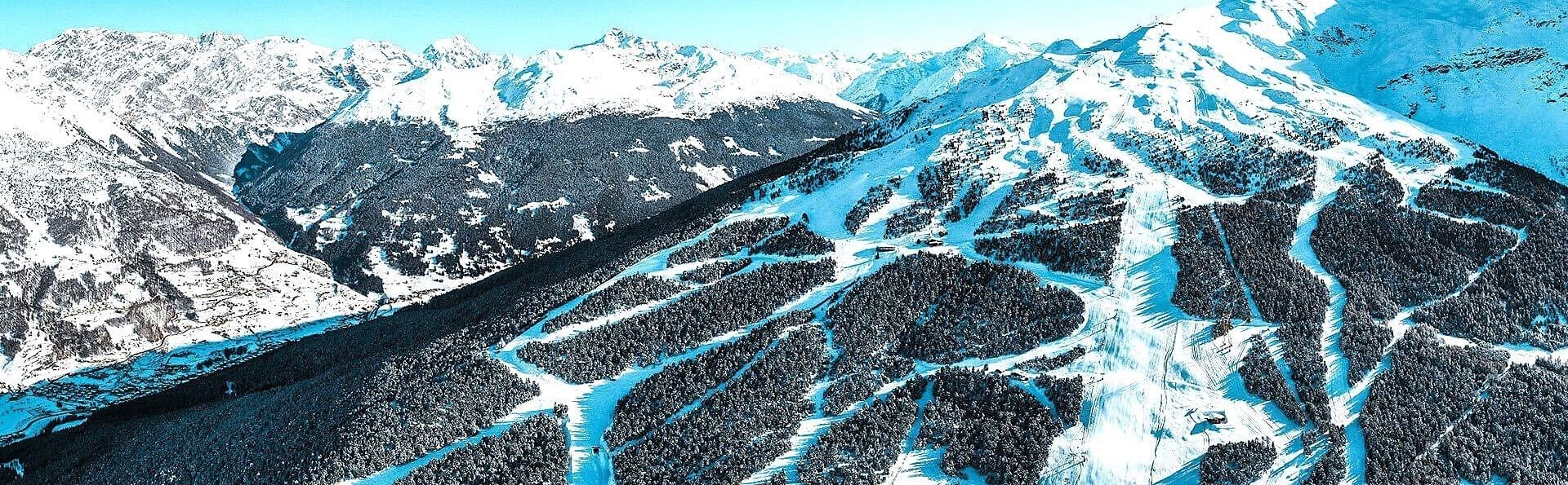 Veduta aerea di una catena montuosa innevata con piste da sci scavate tra fitte foreste sotto un cielo sereno.