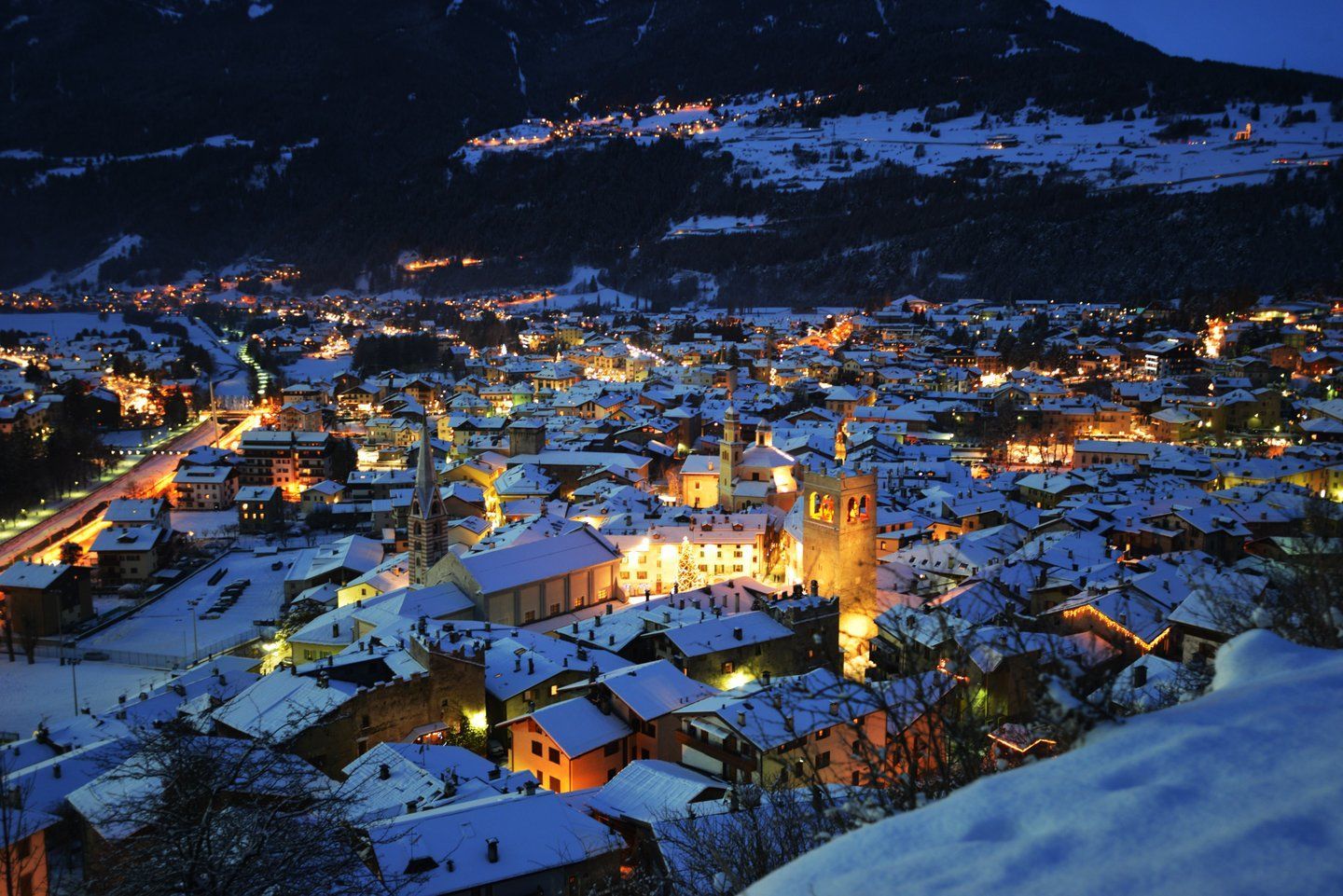 Una città alpina con tetti innevati è illuminata a giorno di notte, incastonata ai piedi di una montagna scura.