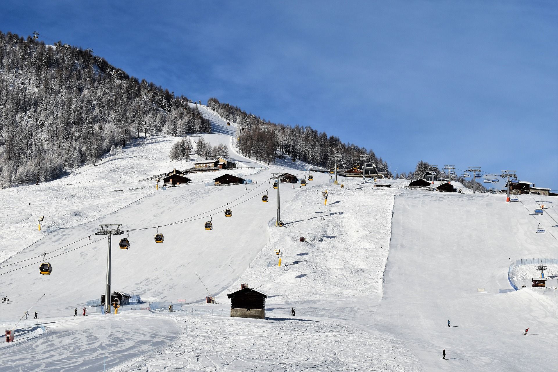 Una località sciistica soleggiata con cabinovie che si muovono su piste innevate, sciatori e chalet in legno sul fianco della montagna.