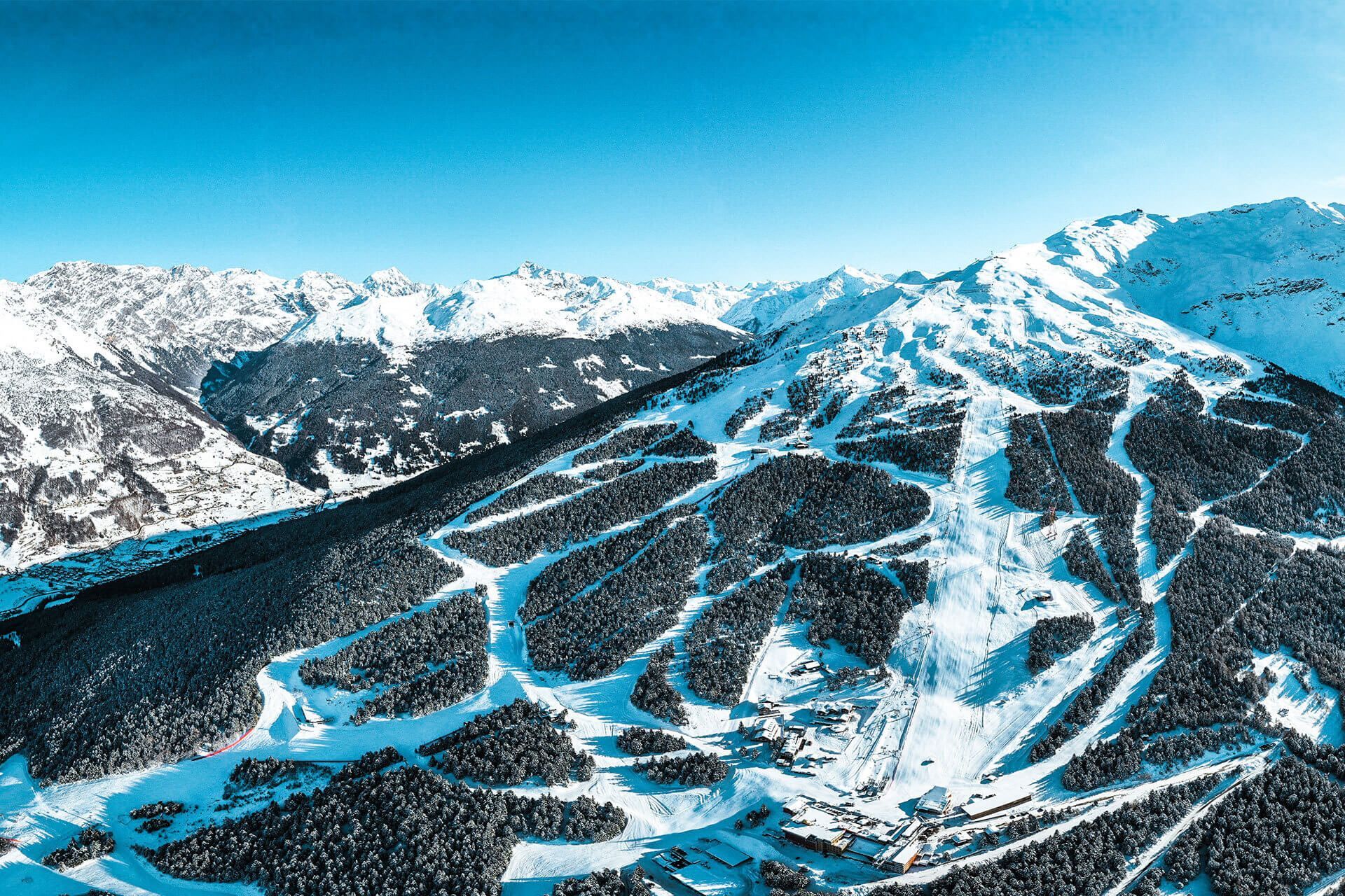 Una vista aerea di una stazione sciistica con diverse piste che attraversano una foresta montana innevata sotto un cielo sereno.