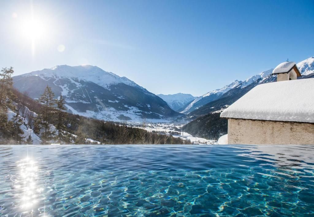 Una piscina a sfioro si affaccia su una valle assolata e montagne innevate, con un tetto innevato in primo piano.