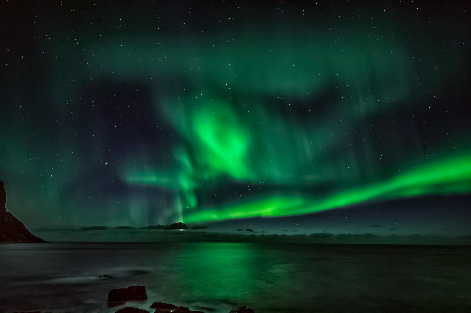L'aurora boreale verde danza nel cielo notturno stellato, riflettendosi sul mare calmo sottostante con rocce in primo piano.