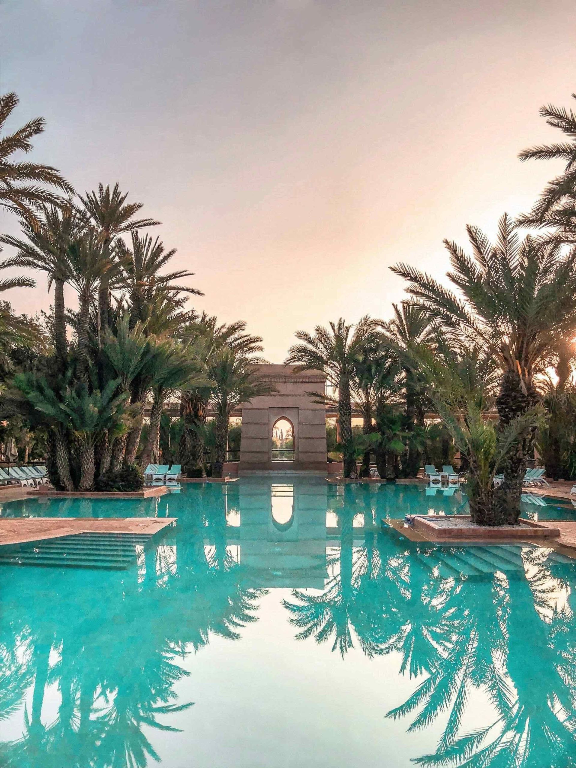 Una grande piscina riflette le palme circostanti e un arco in pietra sotto un cielo al tramonto.