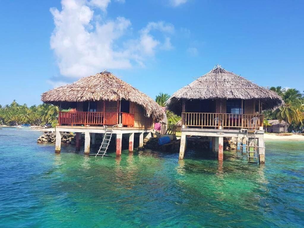 Due bungalow dal tetto in paglia, costruiti su palafitte sopra acque limpide color turchese, con una spiaggia sabbiosa e palme sullo sfondo.