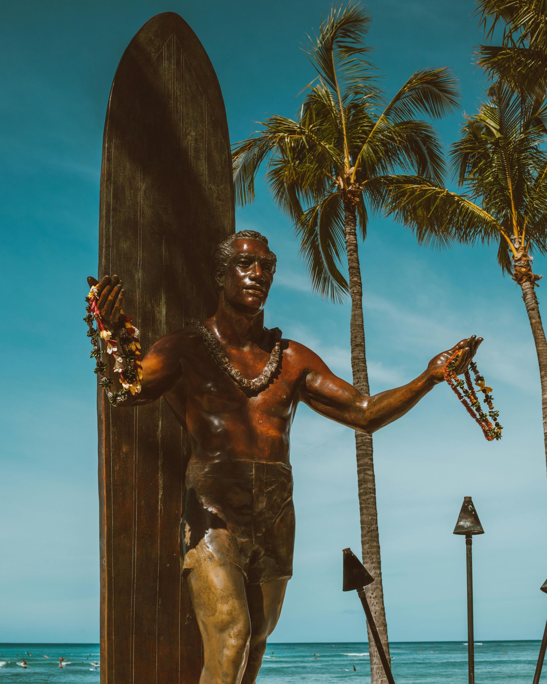 Eine Bronzestatue eines Surfers mit Leis steht neben einem Surfbrett, mit Palmen und dem Ozean im Hintergrund.