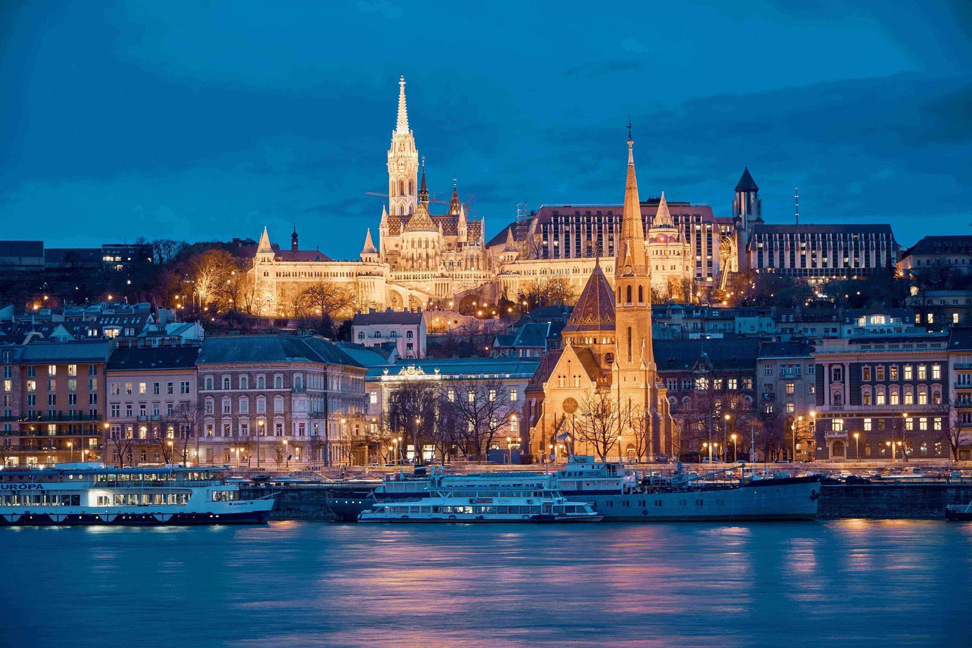 Skyline storico di una città europea con chiese ed edifici illuminati, visto al tramonto da oltre il fiume.