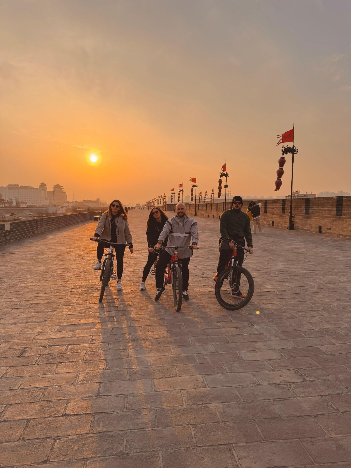 Quattro persone di un gruppo WeRoad in posa con le biciclette su un ampio muro di mattoni della città durante un tramonto nebbioso.