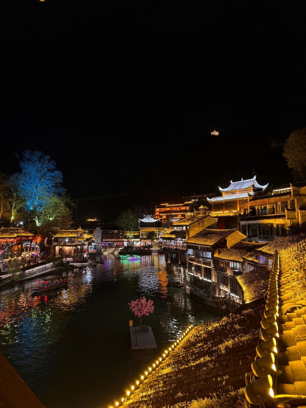 Una cittadina lungo il fiume con architettura tradizionale, illuminata di notte da luci dorate e colorate che si riflettono nell'acqua.