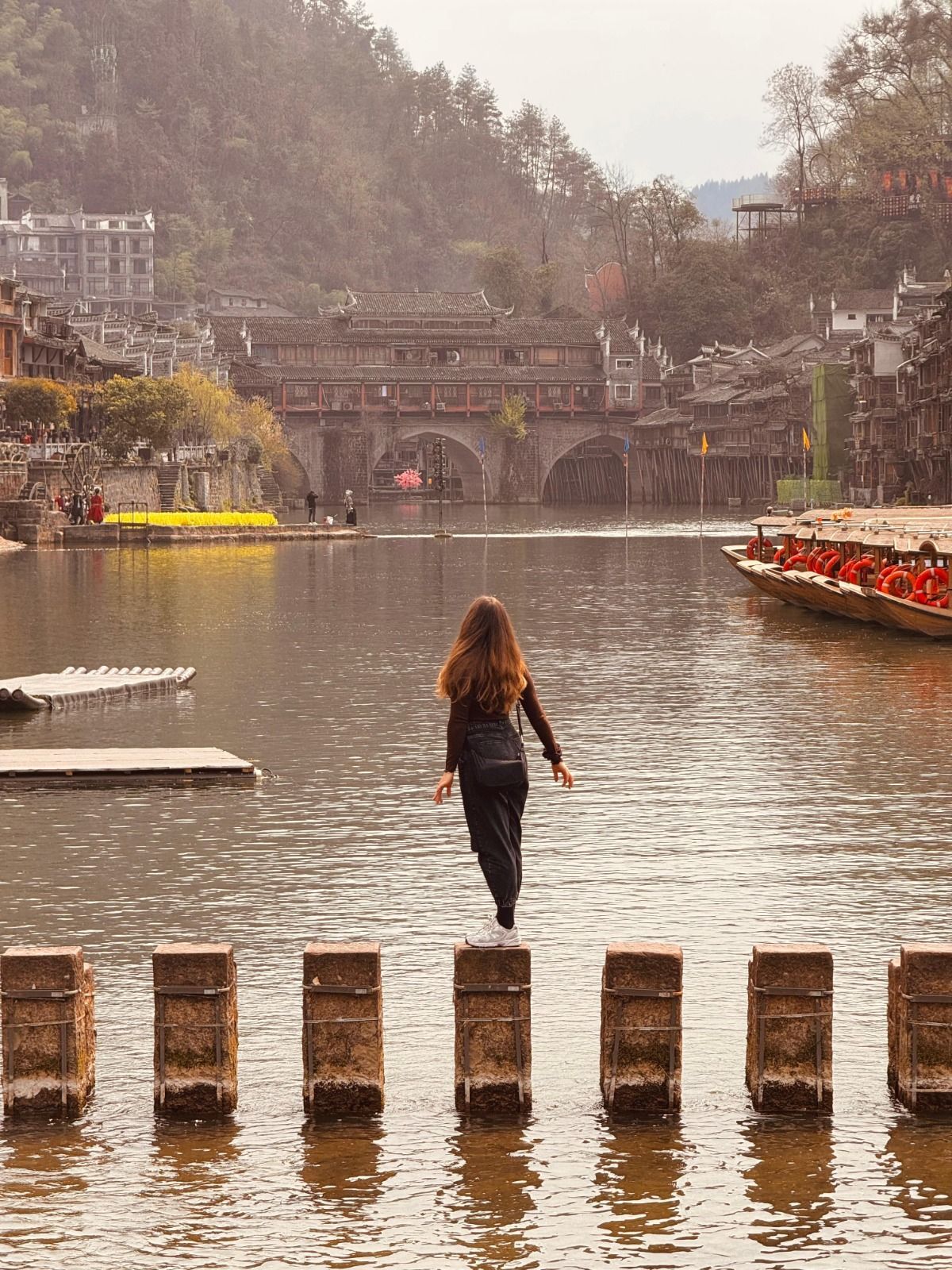 Una donna di spalle alla fotocamera si bilancia su pietre a guado su un fiume, con un villaggio tradizionale e un ponte in lontananza.