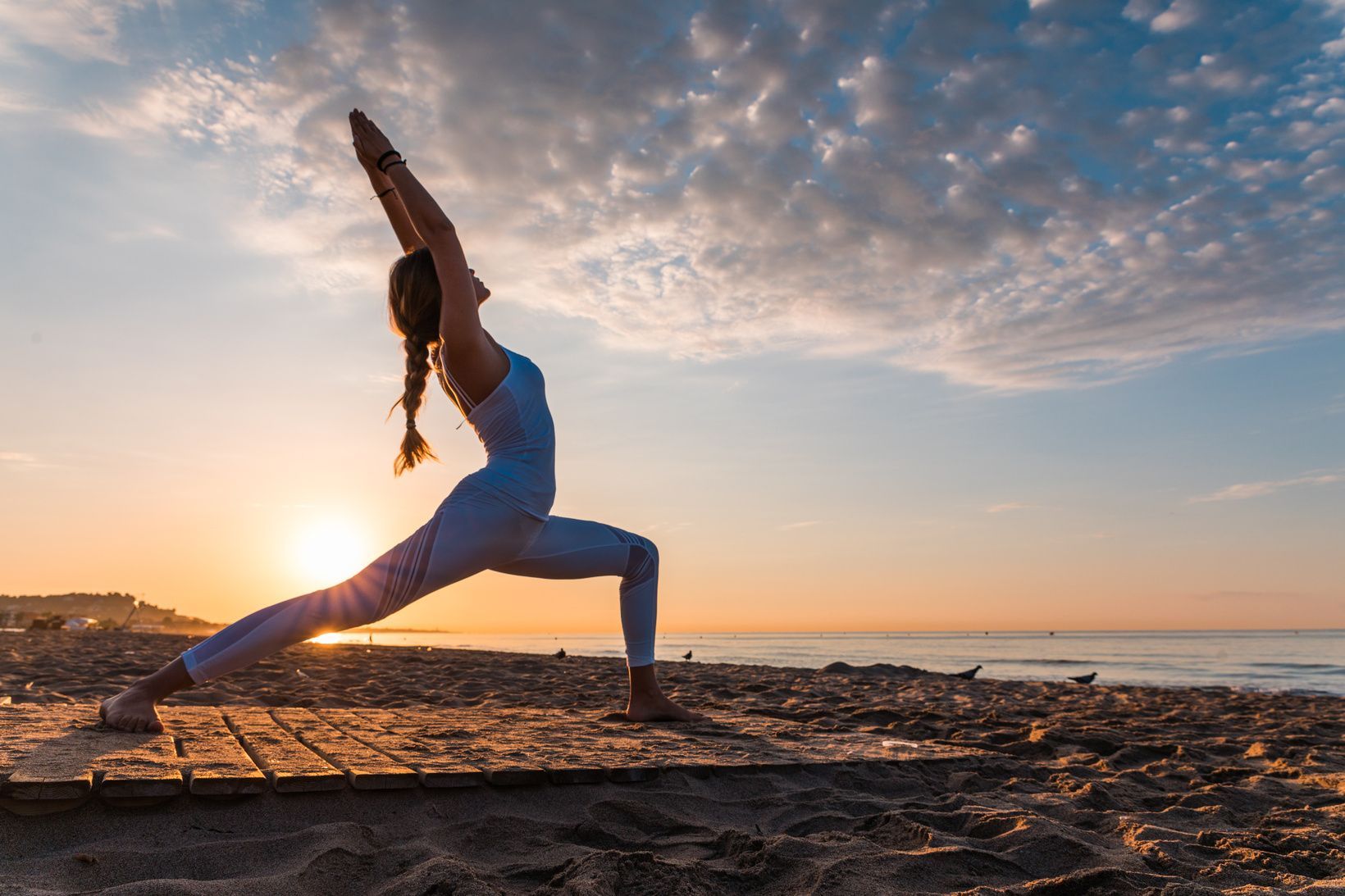 Una donna esegue una posa yoga del guerriero su una piattaforma di legno su una spiaggia sabbiosa all'alba.