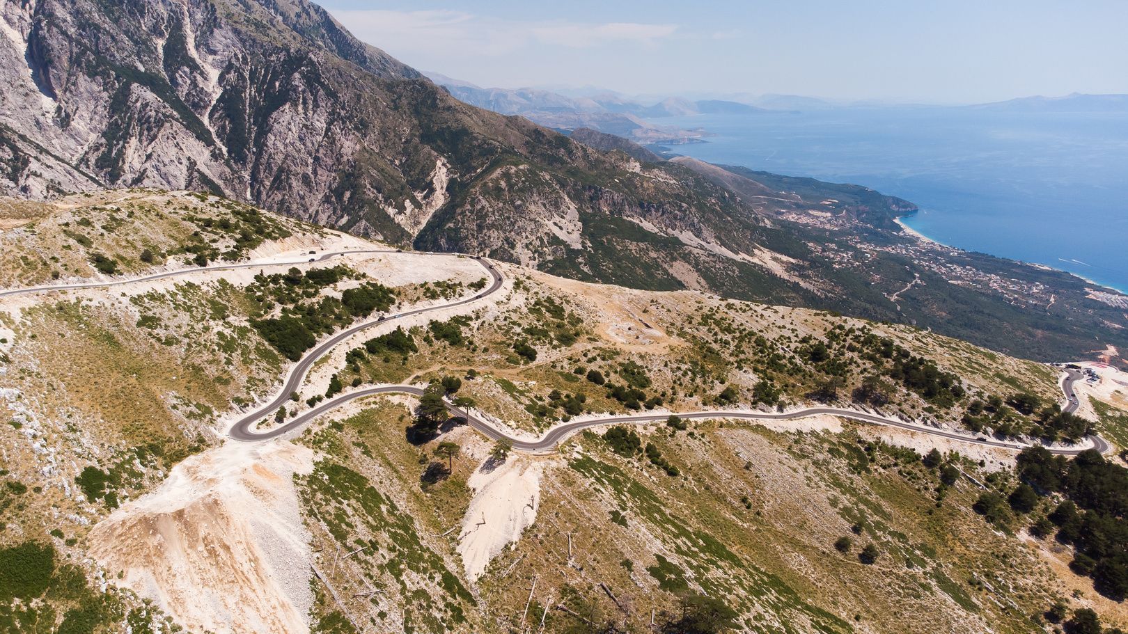 Vue aérienne d'une route sinueuse serpentant à travers un paysage montagneux accidenté, avec la mer bleue et la côte au loin.