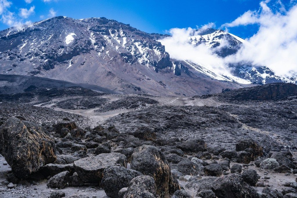 Un paesaggio roccioso e vulcanico con un sentiero tortuoso che conduce verso montagne innevate sotto un cielo azzurro brillante.