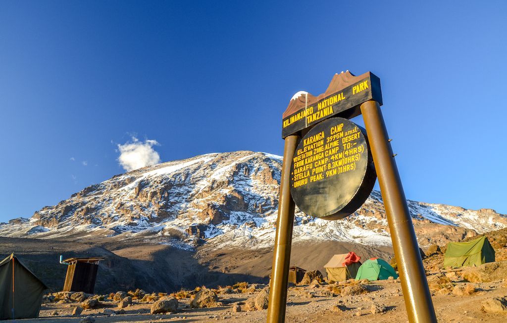 Un cartello del Campo Karanga nel Parco Nazionale del Kilimangiaro davanti a un campeggio, con la vetta innevata della montagna sullo sfondo.